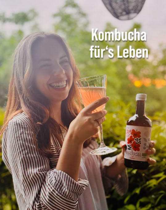 Eine Frau hält ein Glas mit rosa Getränk und eine Flasche Kombucha, im Hintergrund grüne Pflanzen und der Text "Kombucha für's Leben".