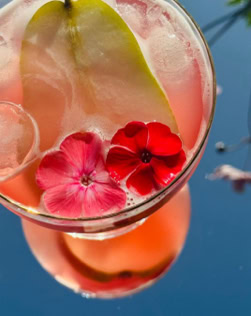 Ein rosa Getränk mit Eiswürfeln, einer Birnenscheibe und zwei roten Blüten in einem Glas.