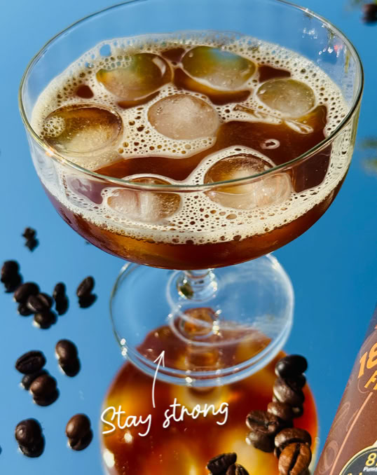 Ein Glas mit einem braunen Getränk und Eiswürfeln, umgeben von Kaffeebohnen auf einer blauen Oberfläche. Darunter steht der Text "Stay strong".