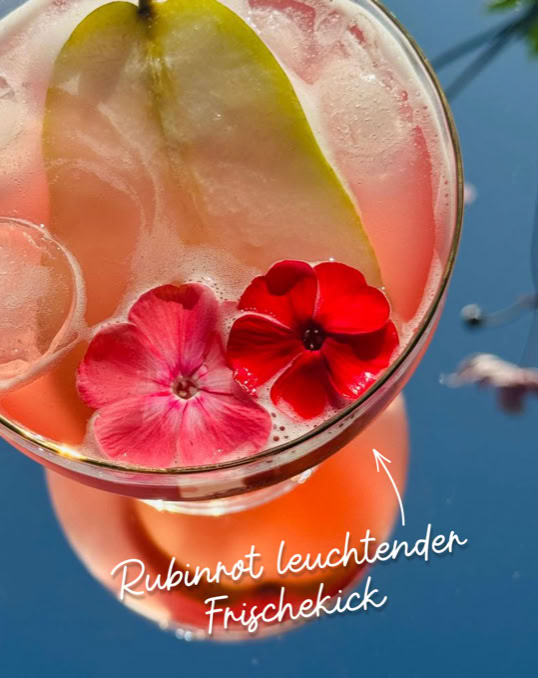 Ein rosa Getränk mit Eiswürfeln, einer Birnenscheibe und roten Blüten, auf einem blauen Hintergrund. Text: "Rubinrot leuchtender Frischekick".