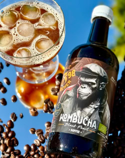 Eine Flasche Kombucha mit einem Schimpansen auf dem Etikett, daneben ein Glas mit schaumiger Flüssigkeit und Kaffeebohnen auf einem blauen Hintergrund.