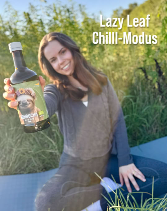 Eine Person sitzt auf einer blauen Matte im Freien und hält eine Flasche mit einem Etikett, das einen Hund zeigt. Im Hintergrund ist hohes Gras zu sehen. Oben rechts steht der Text "Lazy Leaf Chilll-Modus".