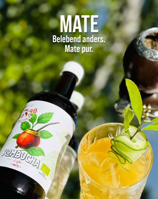 Eine Flasche mit Etikett und ein Glas mit gelbem Getränk und Gurkenscheiben, im Hintergrund unscharfes Grün. Text: "MATE Belebend anders. Mate pur."