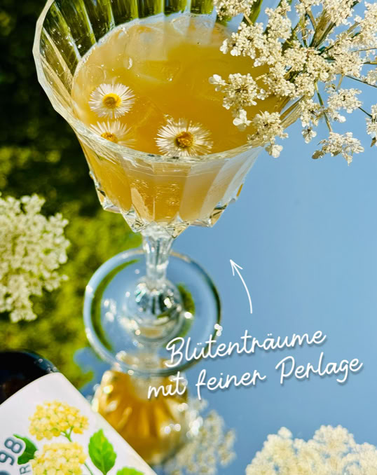 Ein Glas mit gelbem Getränk, dekoriert mit weißen Blüten, steht auf einem Spiegel. Daneben ist eine Flasche mit einem Etikett, das Blumen zeigt.