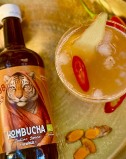 Flasche Kombucha mit Tigeretikett neben einem Glas mit Getränk, Eiswürfeln, Ingwerscheibe und Chilischote, auf einem goldenen Tablett mit Ingwerstücken.