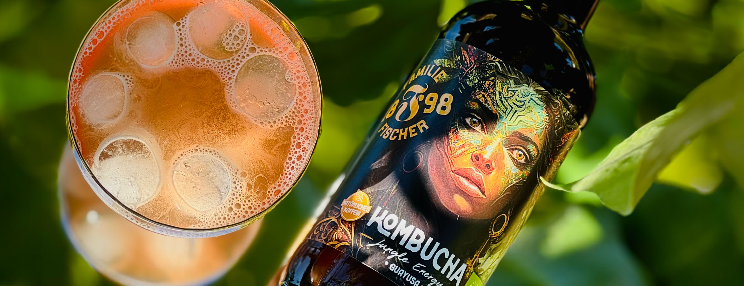 Eine Flasche Kombucha mit einem künstlerischen Etikett neben einem Glas mit einem sprudelnden Getränk, im Hintergrund grüne Blätter.