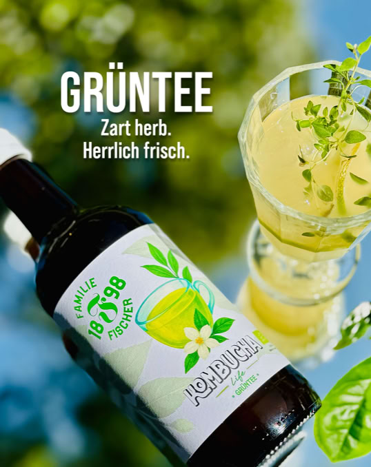 Flasche und Glas mit grünem Tee, umgeben von grünen Blättern, mit Text "GRÜNTEE Zart herb. Herrlich frisch."